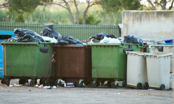 Overflowing trash containers line a street during the daytime.