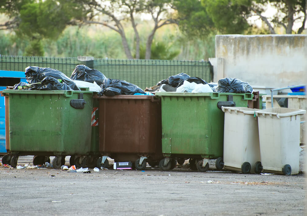 Overflowing trash containers line a street during the daytime.